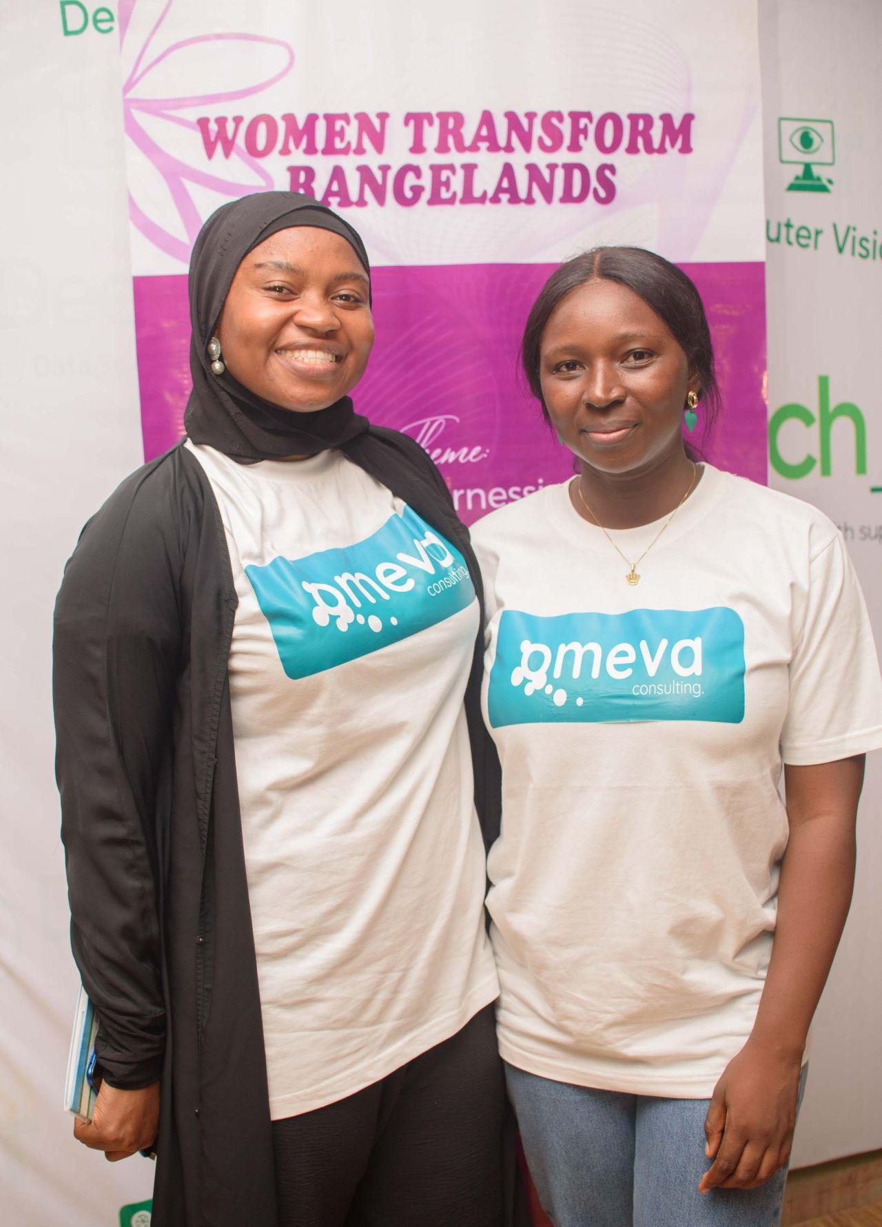Two African women are smiling and standing in front of a banner that reads Women Transform Rangelands