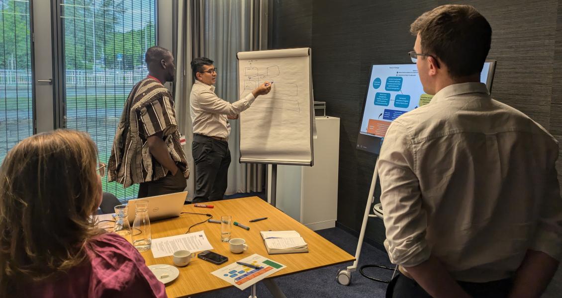 Four people standing look at a person who is presenting information on a flip chart. Two people in the foreground are seen from behind.
